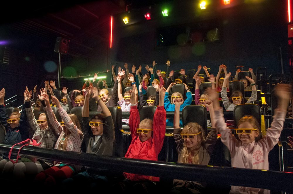 Kinder im Action-Kino mit hochgerissenen Armen und gelben Brillen auf