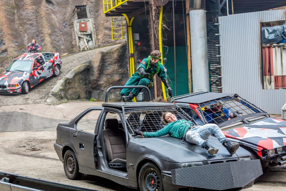 Eine Stuntfrau ist an einem Auto festgebunden, ein Stuntman steht auf dem Autodach