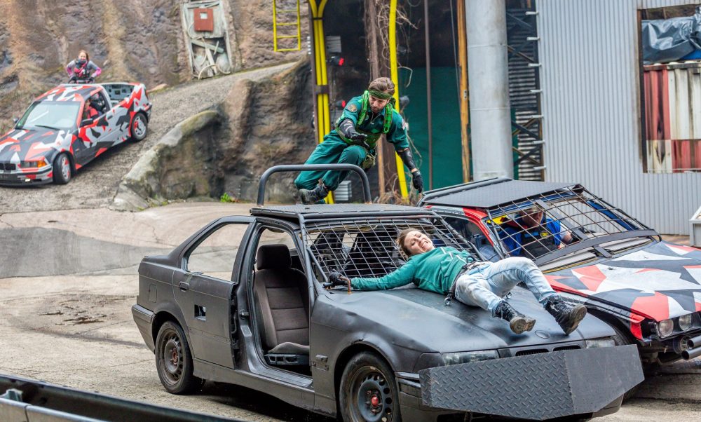 Eine tuntfrau ist an einem Auto festgebunden, ein Stuntman steht auf dem Autodach